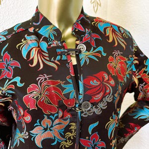 Op de afbeelding: Een zwart jack met een bloemenprint in tinten rood, blauw en goud. Het jack heeft een mandarijnskraag en een knoopsluiting.