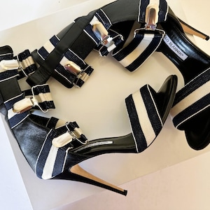 Puede incluir: Par de sandalias de tacón alto a rayas blancas y negras con múltiples correas y hebillas plateadas. Las sandalias tienen punta y tacón de aguja. El nombre de la marca "Manolo Blahnik" es visible en la plantilla.