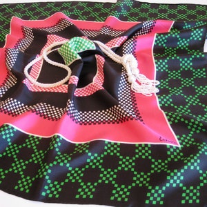 K&ouml;nnte beinhalten: Ein schwarzer, rosa und gr&uuml;ner Seidenschal mit geometrischem Muster aus kleinen Quadraten. Auf dem Schal liegt eine wei&szlig;e Perlenkette.