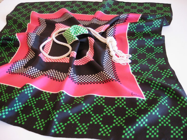K&ouml;nnte beinhalten: Ein schwarzer, rosa und gr&uuml;ner Seidenschal mit geometrischem Muster aus kleinen Quadraten. Auf dem Schal liegt eine wei&szlig;e Perlenkette.