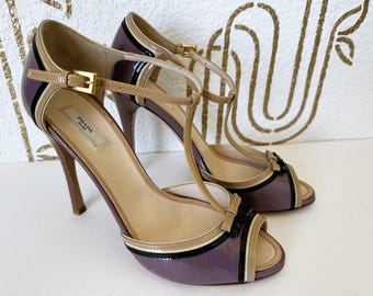 Puede incluir: Un par de sandalias de tacón alto Prada. Los zapatos de punta abierta presentan un acabado brillante de color ciruela con detalles en beige y negro. Las sandalias tienen un diseño de tira en T y cierre de hebilla dorada. El logo de Prada es visible.