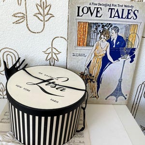 May include: A round, black and white striped hat box with a lid featuring the text 'an exclusive Lisa NEW YORK PARIS' in black script and print. A black hand-shaped sculpture extends from the side, and a black ribbon rests on the lid.