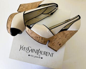 Puede incluir: Par de sandalias de cuña Yves Saint Laurent. Las sandalias presentan una parte superior de tela color crema con ribete negro y un tacón de cuña de corcho. El diseño de punta abierta y la correa del tobillo se suman al estilo de la sandalia. Sobre una caja con el logo de Yves Saint Laurent.