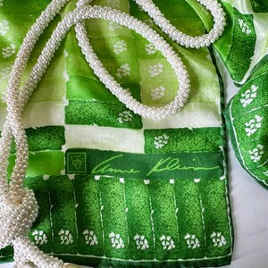 May include: A green and white patterned scarf with a matching section and a white beaded necklace. The scarf features a repeating block design with floral accents and the Anne Klein signature. The necklace is draped across the scarf.