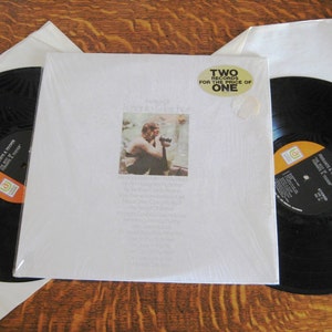 Puede incluir: Un álbum de vinilo con una portada blanca que presenta una fotografía de un hombre. El álbum está sellado en plástico e incluye una pegatina que dice "TWO RECORDS FOR THE PRICE OF ONE". Se ven dos discos de vinilo negro.