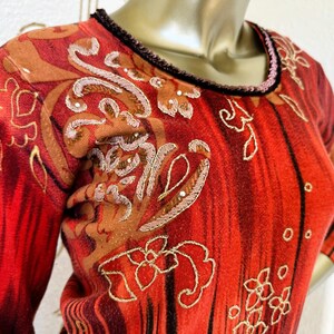 May include: A red sweater with long sleeves, featuring a detailed floral and scrollwork pattern in gold and brown hues. The neckline and cuffs are trimmed in black, with small decorative elements.