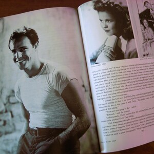 May include: A black and white photograph of a smiling man in a white t-shirt and dark pants, featured in a book. The book is open, displaying the image alongside text. The image is a vintage portrait.