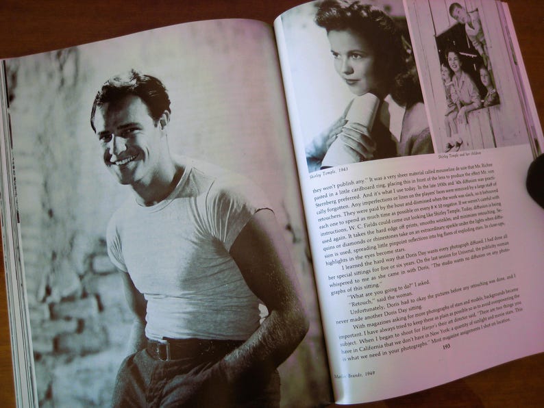 May include: A black and white photograph of a smiling man in a white t-shirt and dark pants, featured in a book. The book is open, displaying the image alongside text. The image is a vintage portrait.