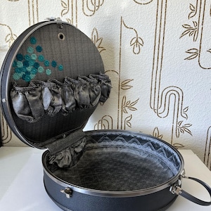 May include: A round black carrying case with a textured exterior and a silver metal handle. The interior is lined with dark gray fabric, featuring a quilted base and a lid with a row of elasticized pockets and teal floral embroidery accents.