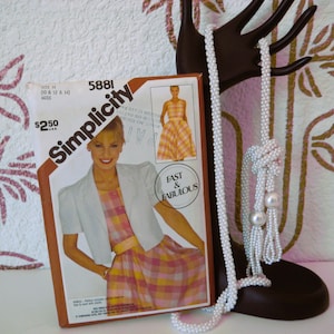 Könnte beinhalten: Eine Vintage-Simplicity-Schnittmusterbox mit einer Frau in einem karierten Kleid und einer Jacke. Eine weiße Perlenkette hängt an einem dekorativen, handförmigen Ständer. Die Schnittmusterbox trägt den Text "Fast & Fabulous".