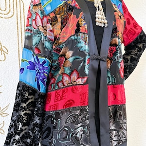 May include: A vibrant patchwork jacket with floral and geometric designs. The jacket has black satin trim and sleeves with a velvet floral pattern. It is open-fronted with a black tie closure.
