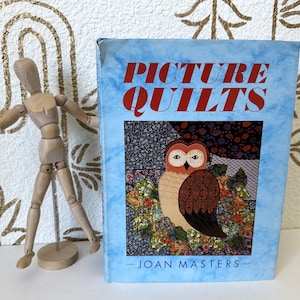 Op de afbeelding: Een hardcover boek getiteld Picture Quilts door Joan Masters met een lichtblauwe gemarmerde kaft met een centrale illustratie van een uil omringd door bloemmotieven, geplaatst naast een kleine houten gearticuleerde kunstenaarspop.