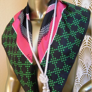 K&ouml;nnte beinhalten: Ein schwarzer und gr&uuml;ner Seidenschal mit einem rosa und wei&szlig;en geometrischen Muster. Der Schal wird um den Hals einer Schaufensterpuppe getragen und hat eine wei&szlig;e Perlenkette mit einem geknoteten Quasten.