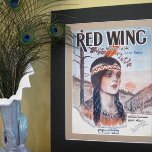 Puede incluir: Una portada de partitura musical vintage enmarcada en negro. La portada presenta una ilustración colorida de una mujer nativa americana con cabello largo y oscuro que lleva una diadema con cuentas y un tocado de plumas. El título "RED WING An Indian Fable Love Song" está impreso en letras rojas y negras.