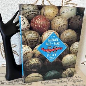 May include: The Baseball Hall of Fame 50th Anniversary Book by Gerald Astor, featuring a cover collage of numerous vintage baseballs with handwritten signatures and historical inscriptions, including a prominent blue diamond-shaped title graphic.