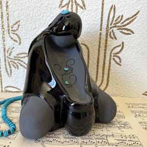Puede incluir: Una escultura de cerámica negra con acabado brillante, adornada con detalles turquesa y un collar de cuentas turquesas. La escultura tiene una forma abstracta única y descansa sobre una hoja de partituras.