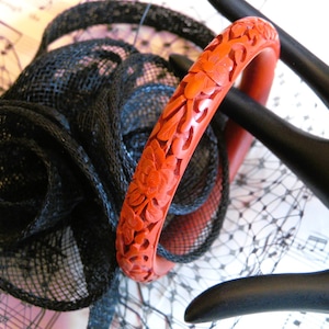 May include: A red carved bangle bracelet with floral designs. The bracelet is on a black lace fabric background.
