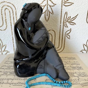 Puede incluir: Una escultura de cerámica negra de una figura sentada, adornada con detalles turquesas. La figura está envuelta en una capa negra brillante, con detalles grises en la ropa. Un collar de cuentas turquesas descansa frente a la escultura.