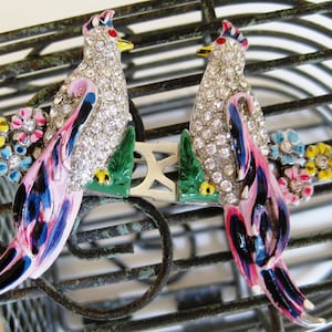 Puede incluir: Un par de broches de pájaro vintage con esmalte colorido y pedrería. Los pájaros están posados en una rama verde con hojas y tienen plumas rosas, azules y negras.