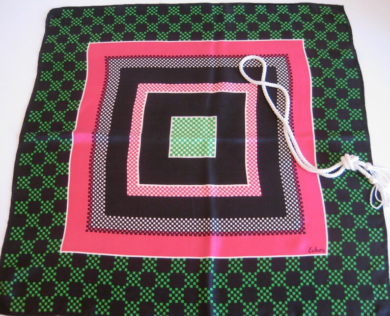 K&ouml;nnte beinhalten: Ein schwarzer Seidenschal mit einem geometrischen Muster aus gr&uuml;nen, wei&szlig;en und rosa Quadraten. Der Schal hat einen wei&szlig;en Quasten und das Wort "Echo" ist in der rechten unteren Ecke aufgedruckt.