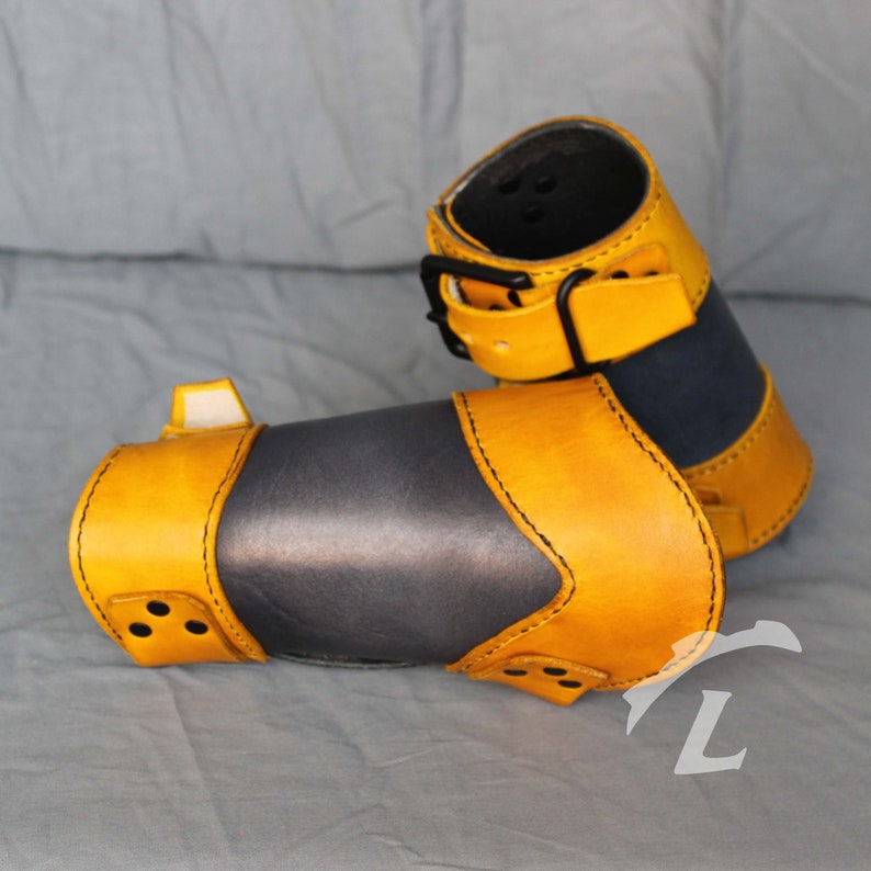 May include: Two yellow leather arm guards with black stitching and a black buckle. The guards are made of two layers of leather, with the top layer being yellow and the bottom layer being black.
