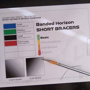 May include: A white sheet of paper with the title "Banded Horizon Short Bracers" and a size chart for small, medium, large, and extra large bracers. The paper also includes a chart for skill levels: basic, intermediate, advanced, and expert. There are two squares, one labeled "2 inch square" and the other labeled "5 cm square". A silver craft knife is lying on the paper.