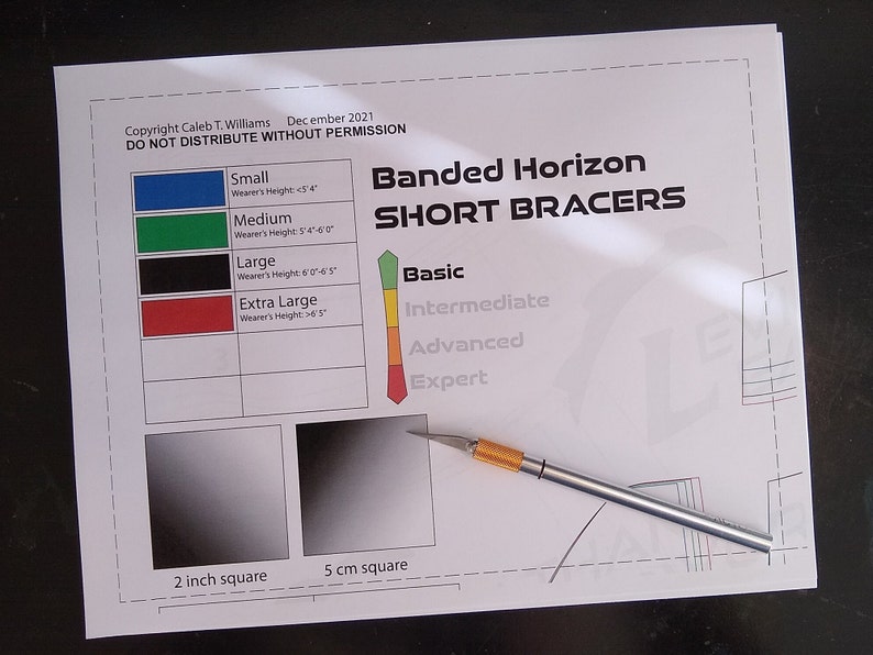 May include: A white sheet of paper with the title "Banded Horizon Short Bracers" and a size chart for small, medium, large, and extra large bracers. The paper also includes a chart for skill levels: basic, intermediate, advanced, and expert. There are two squares, one labeled "2 inch square" and the other labeled "5 cm square". A silver craft knife is lying on the paper.