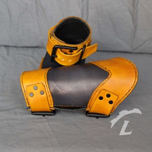 May include: A pair of leather arm guards with a dark blue and tan color scheme. The guards have a strap with a buckle on the top and are secured with a strap and buckle on the bottom.