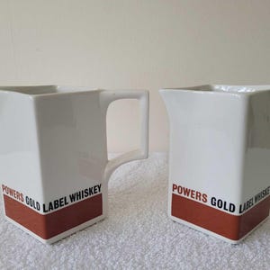 May include: Two white ceramic pitchers with a rectangular shape and a brown band at the base. The text "POWERS GOLD LABEL WHISKEY" is printed on the front. One pitcher has a handle.