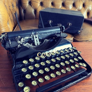 May include: A black Corona typewriter with round, cream-colored keys. The keys are arranged in a QWERTY layout. The typewriter sits on a wooden surface, with a leather couch and a black case in the background. The text "CORONA TYPEWRITER COMPANY Inc." is visible.