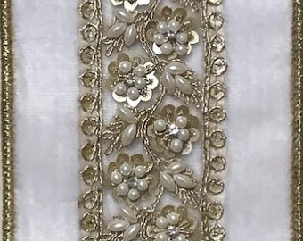 May include: Close-up of decorative trim featuring a central floral design with faux pearls and sequins. The trim is bordered by a gold-colored, beaded edge and is set against a white fabric background. This trim is suitable for embellishing garments or crafts.
