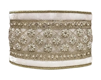 May include: A white, cylindrical decorative object with intricate embellishments. The object features a quilted pattern with floral accents, bordered by gold trim and pearl-like beads. The top and bottom edges are trimmed with gold.