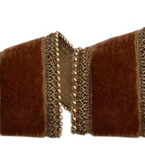 Puede incluir: Primer plano de una cinta decorativa con un centro de terciopelo marrón oscuro y un ribete dorado. El ribete presenta un delicado diseño de cuentas a lo largo de los bordes, añadiendo un toque de elegancia. La cinta está doblada, mostrando su textura y detalles.