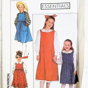 May include: Simplicity Essentials sewing pattern featuring girls' jumper dresses. The pattern includes various styles, such as a blue jumper with pockets, an orange jumper with a white lace trim, and a navy blue polka dot jumper. The pattern size is H (2+3+4).