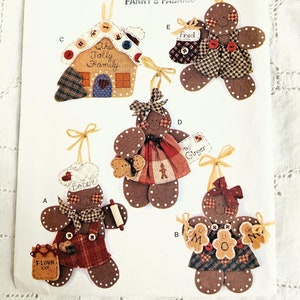 May include: A vintage sewing pattern for making gingerbread ornaments. The pattern includes five different designs: a gingerbread house, a gingerbread person with a rolling pin, a gingerbread person with a bag of flour, a gingerbread person with a sign that says "To Ginger", and a gingerbread person with a sign that says "Mom".
