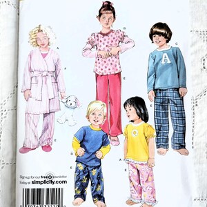 May include: Simplicity sewing pattern number 2826 for children's pyjamas. The pattern includes four different styles: a pink and white striped dressing gown with a matching pyjama set, a pink top with stars and pink trousers, a blue top with a letter A and plaid trousers, and a yellow top with a flower and pink trousers with clouds.