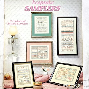 May include: A leaflet titled "keepsake SAMPLERS" featuring nine traditional charted samplers. The samplers are framed and displayed on a pink bedspread. The text includes alphabets, dates, and inspirational quotes.