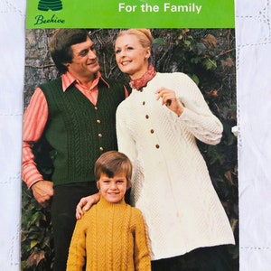 Peut inclure: Couverture de livre vintage avec une famille portant des pulls et des cardigans tricotés. La couverture verte porte le texte "Carefree Arran For the Family" et "Patons Beehive". La famille sourit et pose.