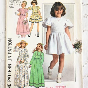 May include: Vintage Simplicity sewing pattern 7954 for girls' dresses. The pattern envelope features illustrations of various dress designs, including a white dress with puff sleeves and a pink gingham dress. The text "ONE PATTERN UN PATRON" is visible.