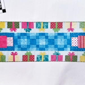 May include: A Party Time table runner featuring a blue checked central panel and colourful gift box motifs. The runner measures 66 cm x 188 cm, ideal for festive or special event decoration.