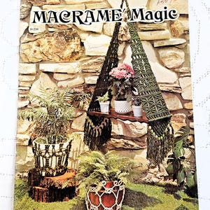 May include: A vintage craft book titled Macrame Magic featuring a cover image of various macrame plant hangers and holders in green and white tones, displaying potted plants on a wooden shelf and tree stumps against a stone wall background.