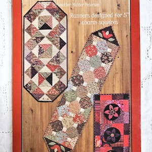 May include: A red cover with the text "Little Charmers" and "Runners designed for 5" charm squares." The cover displays three quilted table runners in various patterns and colors, including floral and geometric designs. The cover also includes the text "Anka's Treasures."