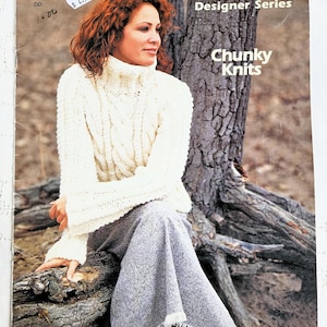 Peut inclure: Un livret de modèles Patons Designer Series Chunky Knits Vol. 2 présentant un pull à col roulé blanc en tricot torsadé avec des manches longues et une jupe grise à ourlet frangé. La couverture affiche le logo et le texte en blanc et noir.