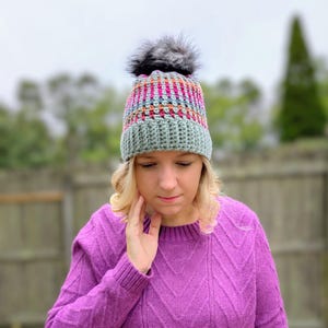 May include: A handmade crochet beanie with a grey faux fur pom-pom. The hat features a ribbed, light green brim and a colourful striped crown in shades of pink, orange, and blue. The hat is designed for cold weather.