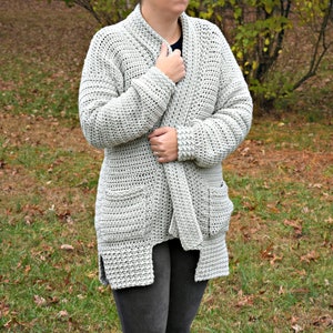 May include: A light gray crocheted cardigan with a shawl collar, pockets, and a tie belt. The cardigan is worn over a black shirt and gray pants.