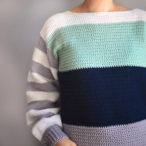 May include: A striped crochet sweater with a white, light green, dark blue, and gray color scheme. The sweater has a boat neck and long sleeves.