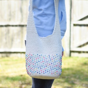 May include: A white crochet tote bag with a colorful beaded design. The bag is worn over the shoulder of a person wearing a light blue shirt and jeans.