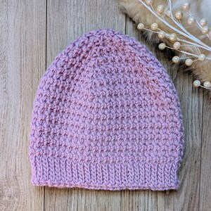 Op de afbeelding: Een lichtpaarse gebreide beanie. De hoed is gemaakt van een textuurgebreide stof en heeft een geribbelde rand. De beanie is effen van kleur en wordt getoond op een houten oppervlak.