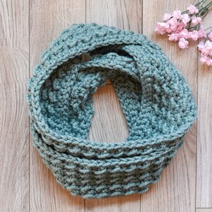 Puede incluir: Una bufanda infinita de crochet azul turquesa. La bufanda está hecha con un hilo grueso y tiene un patrón de punto texturizado.