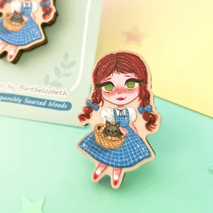 Peut inclure: Épingle en bois représentant une illustration de dessin animé d'une fille en robe bleue tenant un panier avec un chat noir. L'épingle a des cheveux roux et des yeux verts. L'épingle est sur une carte vert clair avec du texte.